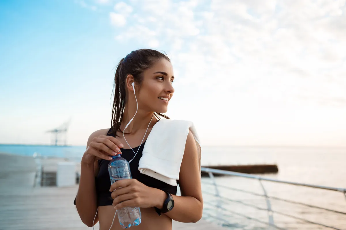 Woman by the sea after exercise holding a water bottle and a towel